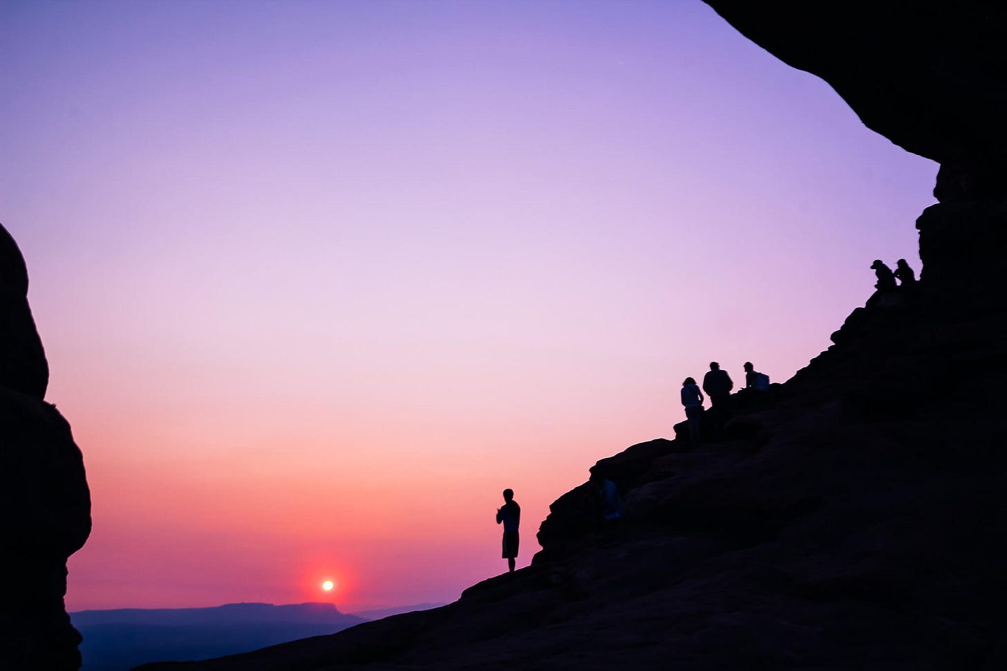 Photo of a Sunrise at Arches National Park taken around 5am a few summers ago by Claire, through an opening in the rocks with people sitting on them looking at the sunrise, rocks and people are in the shadows enhancing the contrast with the sunrise.