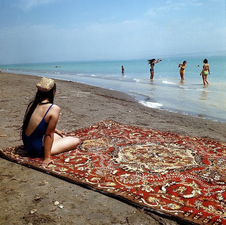 This may contain: a woman sitting on top of a rug next to the ocean while others play in the water