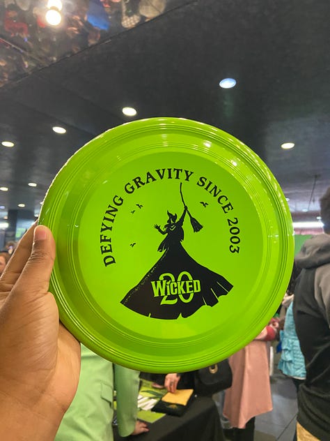 From top left to bottom right - going across horizontally. Image 1) Alexa is holding up a green frisbee that says Defying Gravity since 2003 in black font. The text is curved around the frisbee, in a half circle. In the center, there is a black stenciled picture of Elphaba holding up her Broome. In the middle of skirt, the text “Wicked” is printed with a 20 in the background of the word. Both ‘Wicked’ and the number ’20’ are highlighted in green font. Image 2) The back of Alexa’s left hand. She has a temporary tattoo printed that reads “Toss Toss” in red cursive ink. The two T’s are connected, and there is an asterix/flower type symbol over the second to last S. In smaller font, “Wicked” is printed in all caps. Image 3) Alexa is posing in front of a replica of the clocktower, as seen in the Wicked production. At the top of the clock tower, which is wooden, with antique looking wheels and roman numerals, there is a dragon neck (with its head cut off in the photo) with webbed looking wings. Alexa posing side wise, with a surprised expression on her face, as if she is being scared by someone. She is wearing a polka dotted scarf in her hair, and a pink collared shirt (you can only see her from the shoulders up.) Image 4) Alexa is posing with Carrie St. Louis, who played Glinda on Broadway! Carrie is on the left, wearing a pink satin looking pant suit with a tie around the blazer. Her blond hair falls around her collar bone. She is smiling. Alexa is wearing the same outfit as she was in the previous photo. You can also see her jeans in this photo, along with a pink pin on her shirt. Carrie and Alexa are standing in front of pink background, blocking text that says “it’s good to see me, isn’t it?” Image 5) Alexa is holding up a pink gift bag that reads “Let’s have a celebration the Glinda Way” and the Wicked 20 logo. You can see two pink satin scrounges and a bracelet with pink glass beads on her wrist. You can also make out a few audience members in back of the bag. Image 6) Alexa is holding a light pink baseball cap with the Wicked 20 logo on the front. The text, Wicked, is in White, while the 20 is in green. The entire logo is outlined in black. You can see audience members again, and a little bit of the stage. Image 7) Alexa is holding a pin with the Wicked 20 logo. Wicked is in white, and 20 is in green. Both are outlined in gold. The base of the pink is a golden color, and the outline of the pin has roman numerals like a clock face - in dark green. Image 8) The back of two women, wearing two of Glinda’s iconic costumes - the pink, strapless, frilly, sparkly, short dress from Dancing through Life (left) and the blue dress from the top of the show (right) The blue dress is longer for the show, but this woman’s dress is a bit shorter.