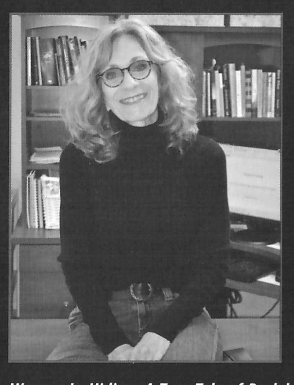 A smiling woman with glasses and curly hair, wearing a black sweater, sits in an office with bookshelves, conveying a friendly mood.