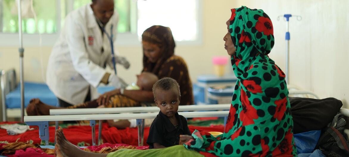 A woman in a green and red patterned headscarf sits on a hospital bed with a young child in Dollow, Somalia, while medical staff treat other patients in the background. A woman in a green and red patterned headscarf sits on a hospital bed with a young child in Dollow, Somalia, while medical staff treat other patients in the background.