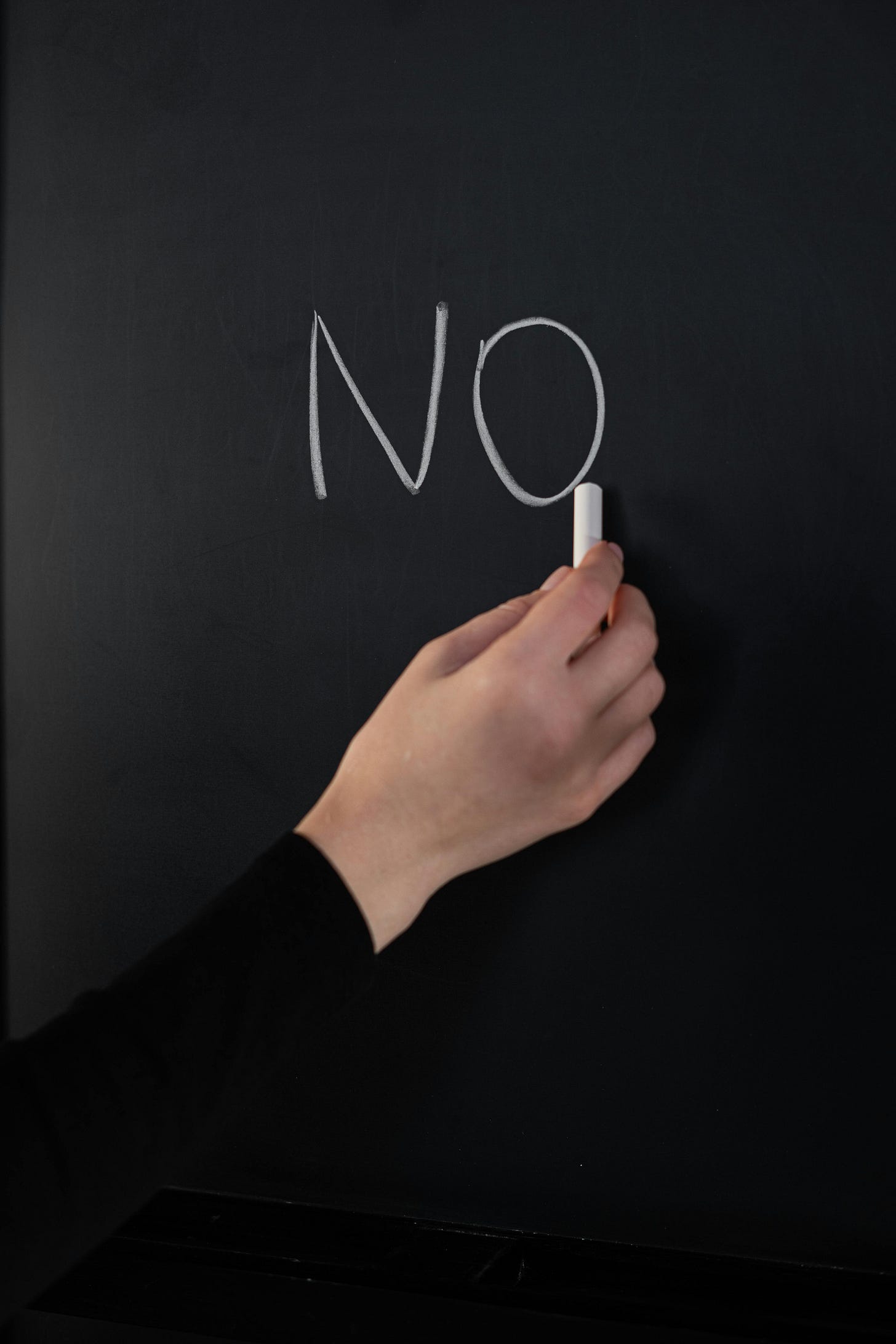 A person with a white hand and black long-sleeved shirt writes the word "NO" on a black chalkboard in white chalk