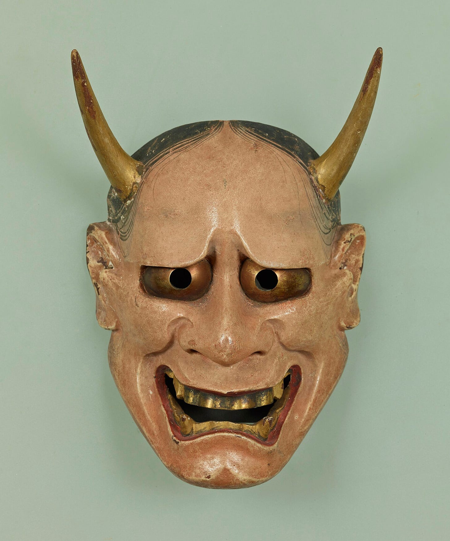 A carved wooden mask of a horned demon woman with golden eyes, a wide grinning mouth full of teeth and fangs, and strands of black hair painted across a pale forehead. A carved wooden mask of a horned demon woman with golden eyes, a wide grinning mouth full of teeth and fangs, and strands of black hair painted across a pale forehead.