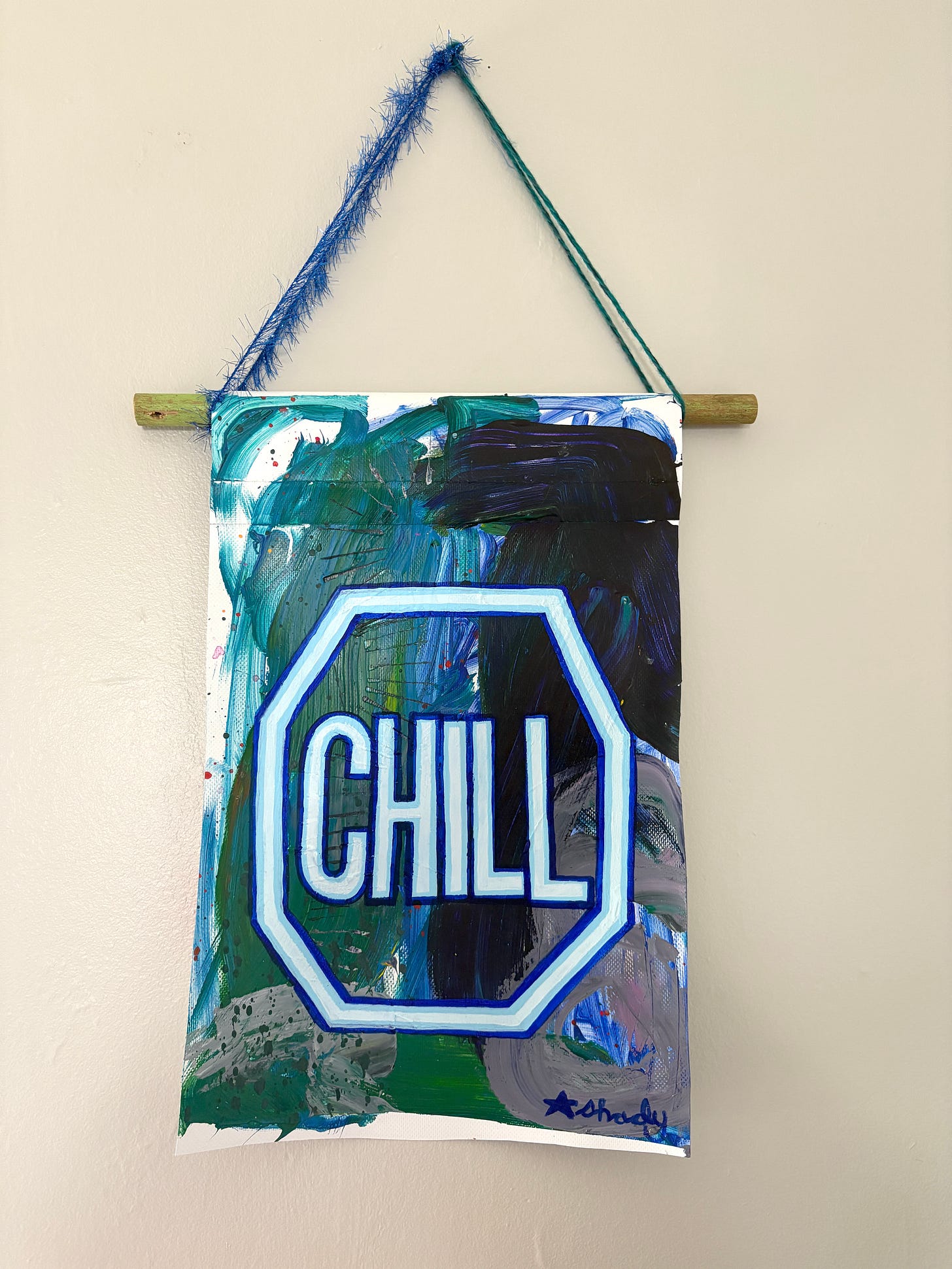 Small painted banner hanging on a wall that says "chill" inside a stop sign shape on an abstract background. The color scheme is blue, green, and grey. 