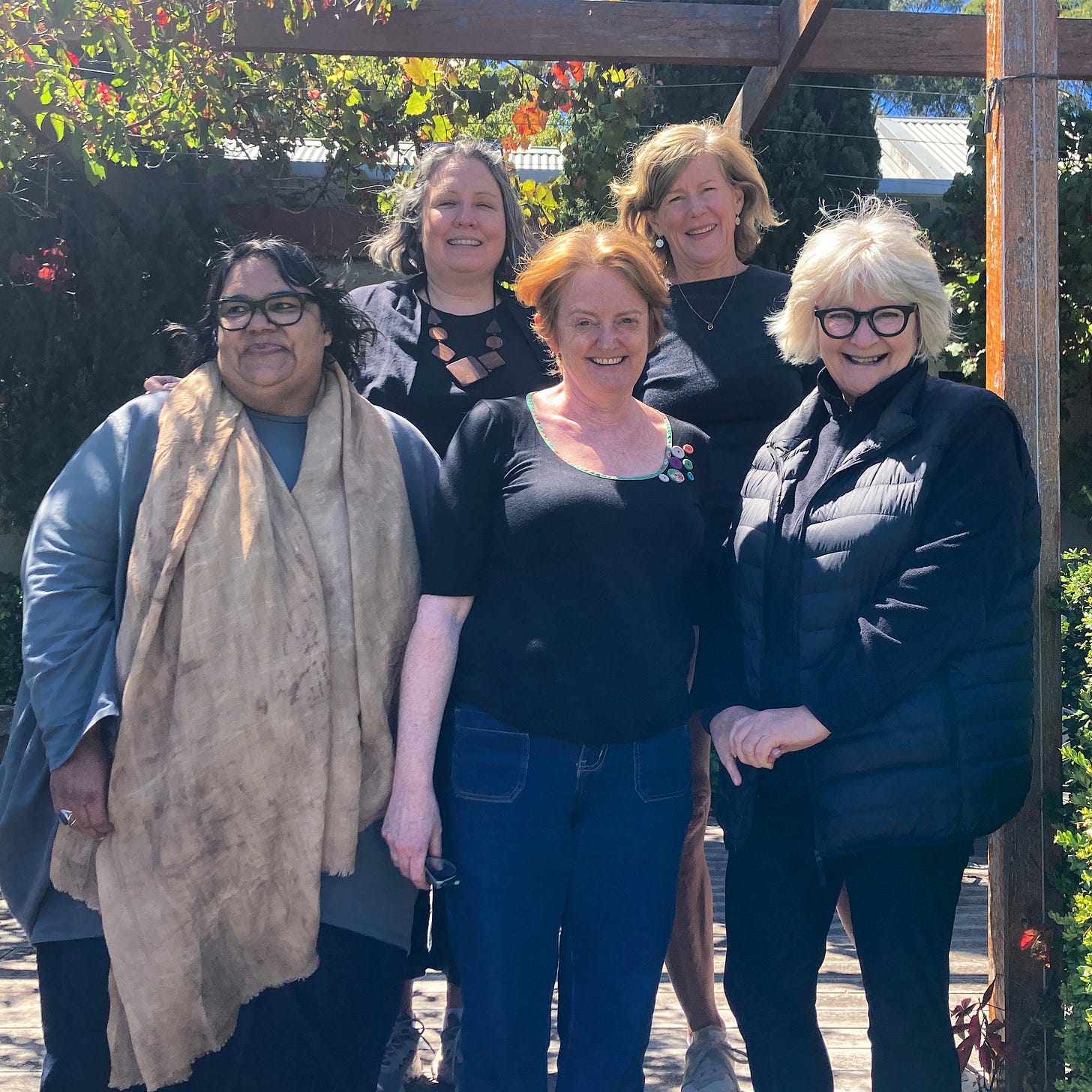 Five women smiling and standing on the edge of a veranda with a vine