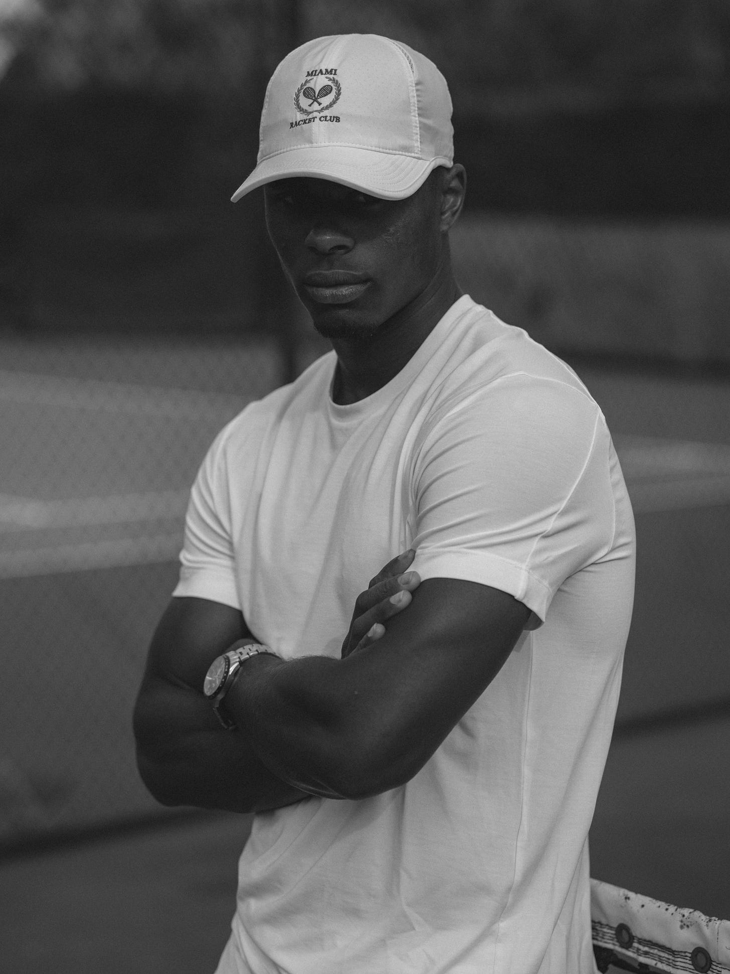 Black and white image of Charles Bertimon posing in a white Jaques athletic outfit and Miami Racket Club hat. He stands with his hands folded, expression serious, embodying calm confidence without a racket.