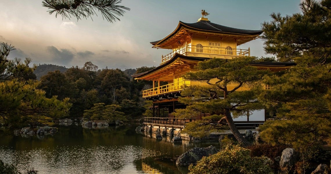 Kinkaku-ji (the Golden Pavilion) in Kyoto, Japan, glowing warmly at golden hour. The iconic three-storey Zen Buddhist temple is covered in gold leaf and reflected in the still waters of Kyoko-chi (Mirror Pond) below. Surrounding the pavilion are traditional Japanese pine trees and carefully placed rocks, with forested hills and a dramatic cloudy sky in the background.