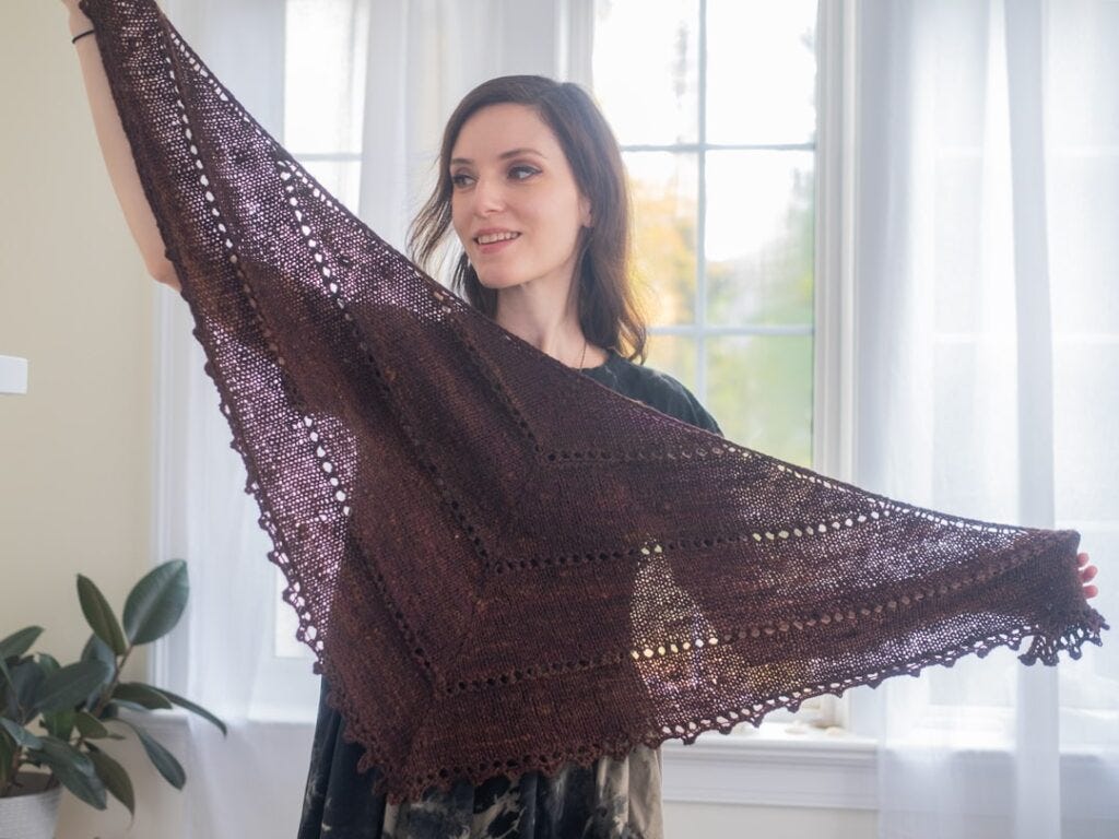 Kristin Lehrer proudly displays her completed Age of Brass and Steam Shawl, hand-knit in a rich chocolate hue. The shawl features an openwork lace with picot edge and a triangular shape. The subtle sheen of the yarn catches the light, highlighting the meticulous stitch work and the cozy yet airy quality of the piece, perfect for adding a touch of warmth and style to any outfit. Kristin Lehrer proudly displays her completed Age of Brass and Steam Shawl, hand-knit in a rich chocolate hue. The shawl features an openwork lace with picot edge and a triangular shape. The subtle sheen of the yarn catches the light, highlighting the meticulous stitch work and the cozy yet airy quality of the piece, perfect for adding a touch of warmth and style to any outfit.