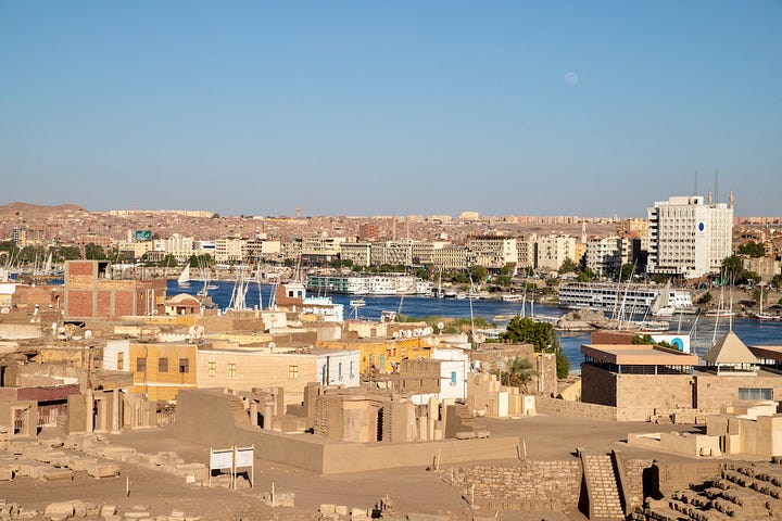 Top left: A busy market street in Aswan with vendors and shoppers beneath a mosque minaret and tangled overhead wires Top right: Two women sitting together sharing a phone beside a table of cosmetics and bottled goods, with a woman in a niqab seated nearby Bottom right: An elevated view across the rooftops of old Aswan toward the Nile, with feluccas and cruise ships moored along the banks and the modern city beyond Bottom left: A young man crouched over an open telecommunications junction box with tangled colored wires, while a woman in a pink hijab and a shopkeeper stand in the doorway behind him