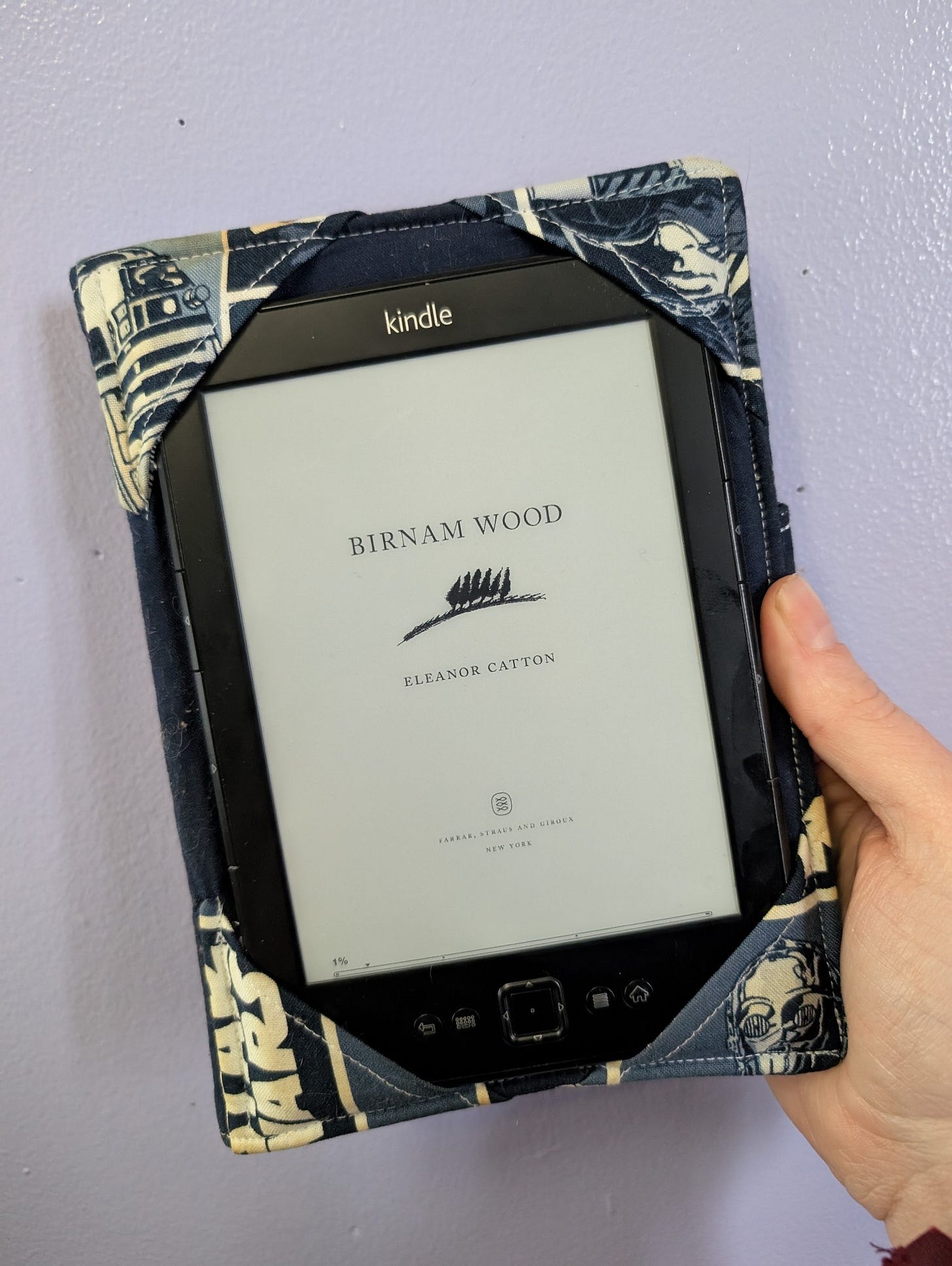 a kindle being held up against a purple background, the screen displaying "Birnam Wood" and author Eleanor Catton