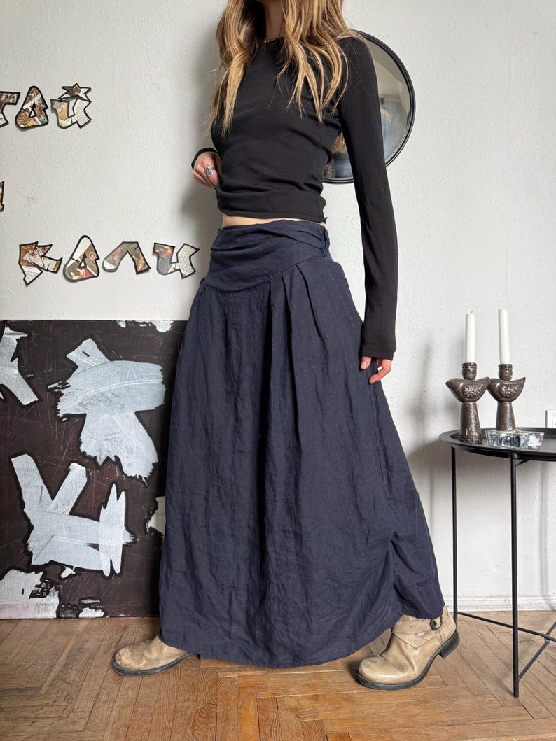 May include: A long-sleeved black top paired with a navy blue maxi skirt. The skirt features a high waist and a flowing, pleated design. The outfit is completed with tan ankle boots. The background includes abstract art and a small side table.