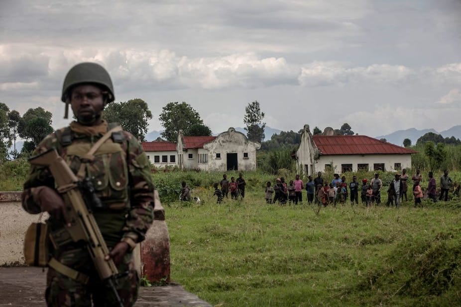 A male soldier holding a gun while a group of children line up behind him in front of white washed buildings that stand on a large grassy field A male soldier holding a gun while a group of children line up behind him in front of white washed buildings that stand on a large grassy field
