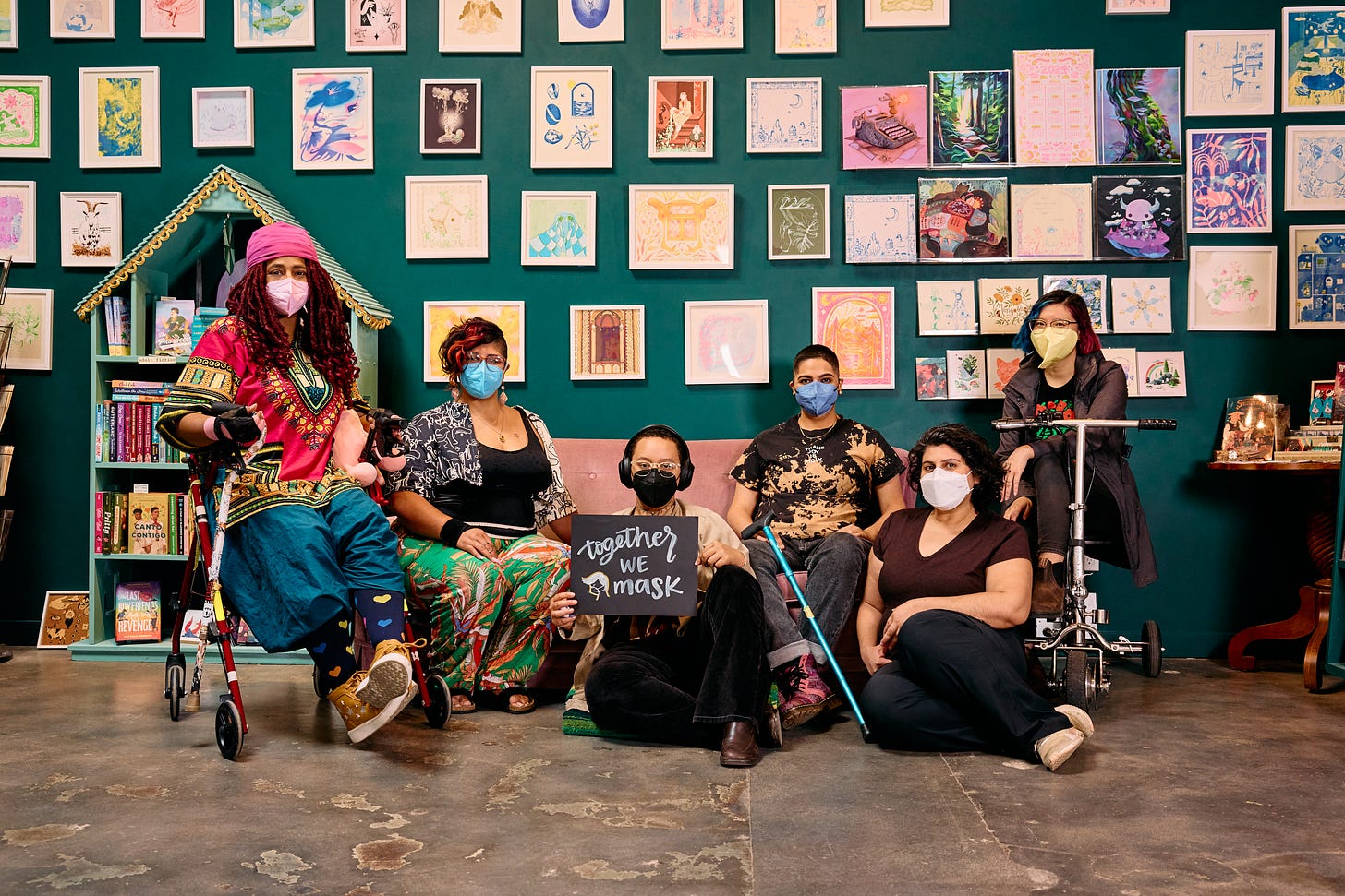 Six disabled people of color sit inside an art-filled bookstore, wearing a mix of KN-95 and N-95 masks in different shades.  In the center, a Black person sits in front of a couch, wearing glasses and noise-canceling headphones, while holding up a sign reading, “Together We Mask.”  On their left, a Black person rests on their rollator and an Indigenous woman sits on the couch. On their right, a Latinx person sits on the couch with their cane resting against their legs, a Latinx woman sits on the floor, and an Asian person relaxes in their mobility scooter.