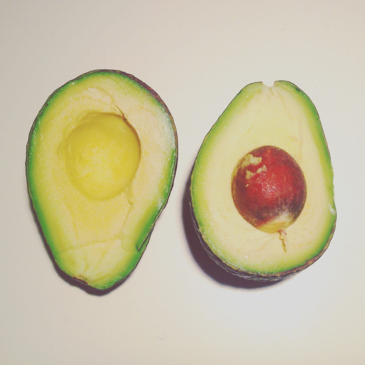 An avocado that has been cut in half sits on a white table. The seed sits in the right half. An avocado that has been cut in half sits on a white table. The seed sits in the right half.