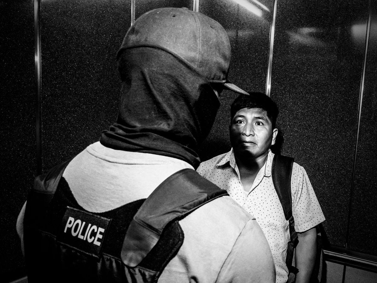 A black-and-white photo of a federal agent, who is wearing a police vest, looming over another man.