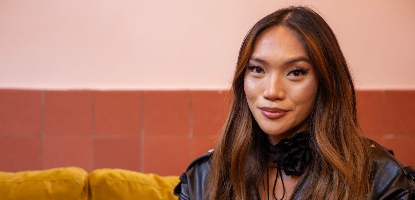 A lady with long hair and a flawless finish of make-up sits on a yellow sofa. She is wearing a black leather jacket.