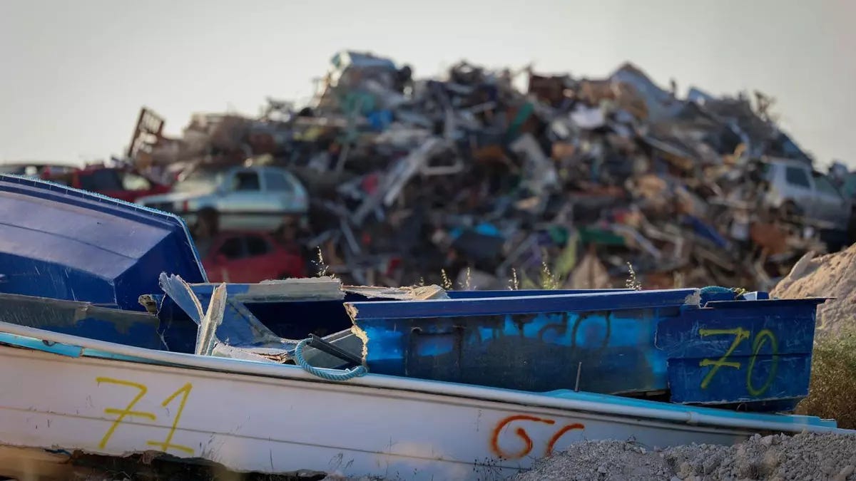 In primo piano si vede il relitto di una barca blu e bianca, danneggiata e con numeri scritti a vernice sullo scafo. Sullo sfondo, fuori fuoco, si erge una massiccia collina di rottami, composta da parti di imbarcazioni, metallo e altri detriti accumulati, sotto un cielo chiaro. In primo piano si vede il relitto di una barca blu e bianca, danneggiata e con numeri scritti a vernice sullo scafo. Sullo sfondo, fuori fuoco, si erge una massiccia collina di rottami, composta da parti di imbarcazioni, metallo e altri detriti accumulati, sotto un cielo chiaro.