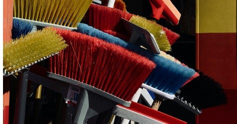 Coloured photo of multi-coloured brooms standing in a pile with broom heads facing up.