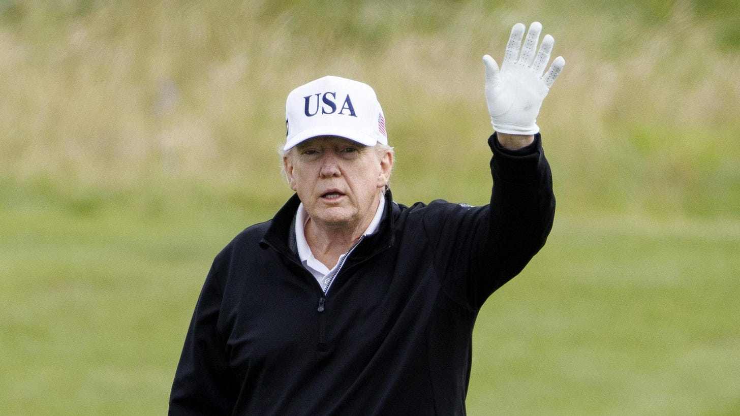 Donald Trump waves a white-gloved hand as he walks on his Turnberry golf course. He is waring a black jumper and a white USA hat