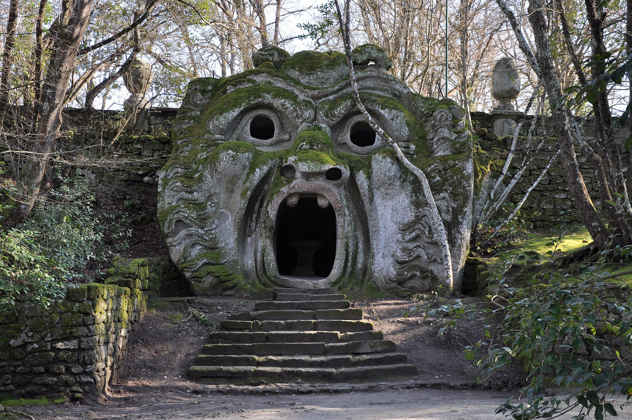 A large stone sculpture of a monstrous face with an open mouth serving as an entrance, covered in moss and surrounded by trees. Located in the Park of the Monsters in Bomarzo, Italy.