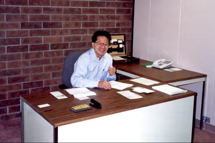 Young Jensen Huang seated at a desk with papers in an office setting.