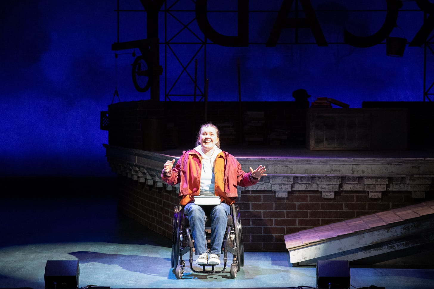 production photo of Meredith Aleigha Wells, a wheelchair user smiling and singing centerstage, hands slightly extended out to the sides.