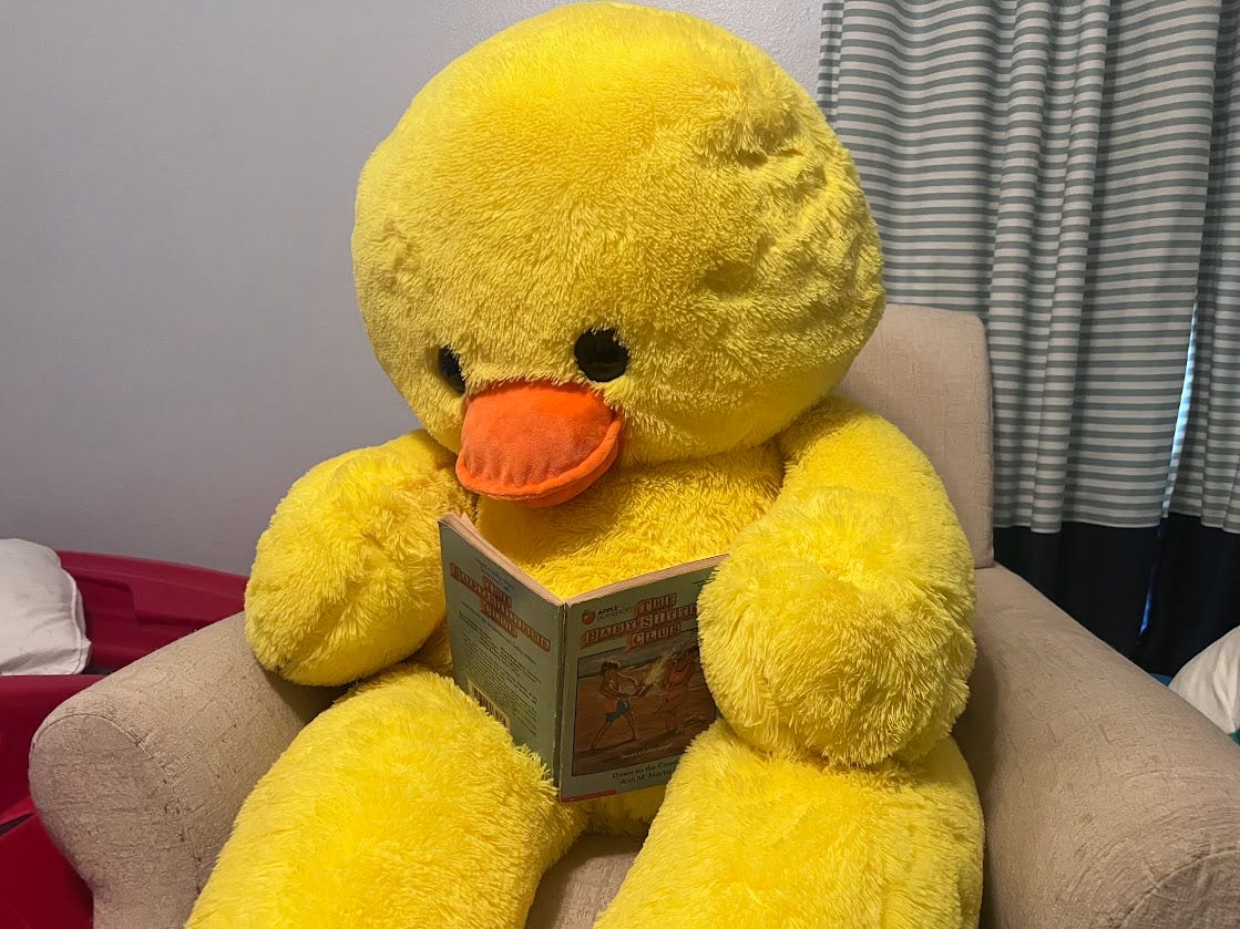 Photo of a giant stuffed duck sitting in a rocking chair reading a Baby-sitters Club book