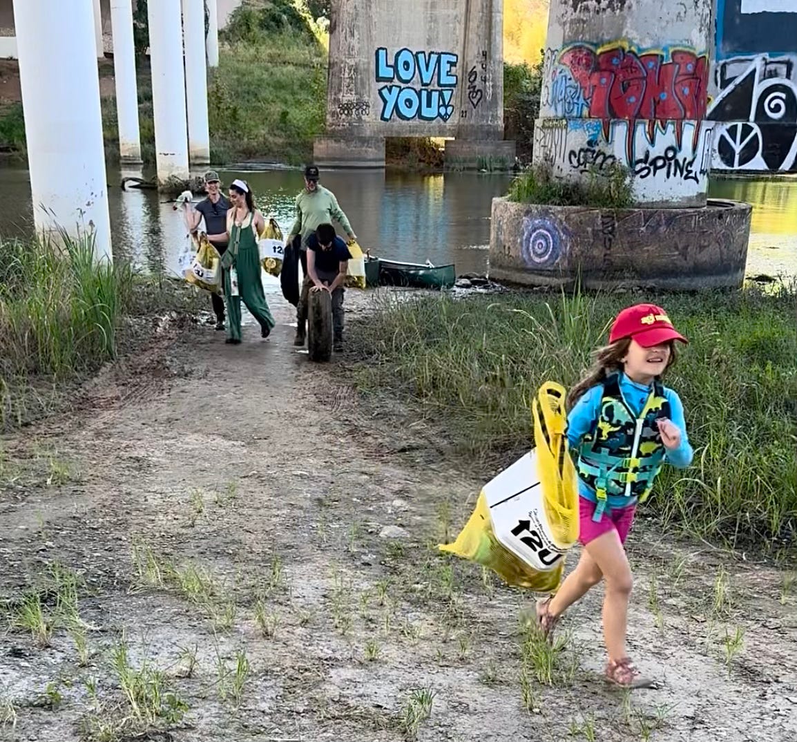 6-year-old girl in foreground and four young adults in background, all hauling big bags of trash collected from an urban river.
