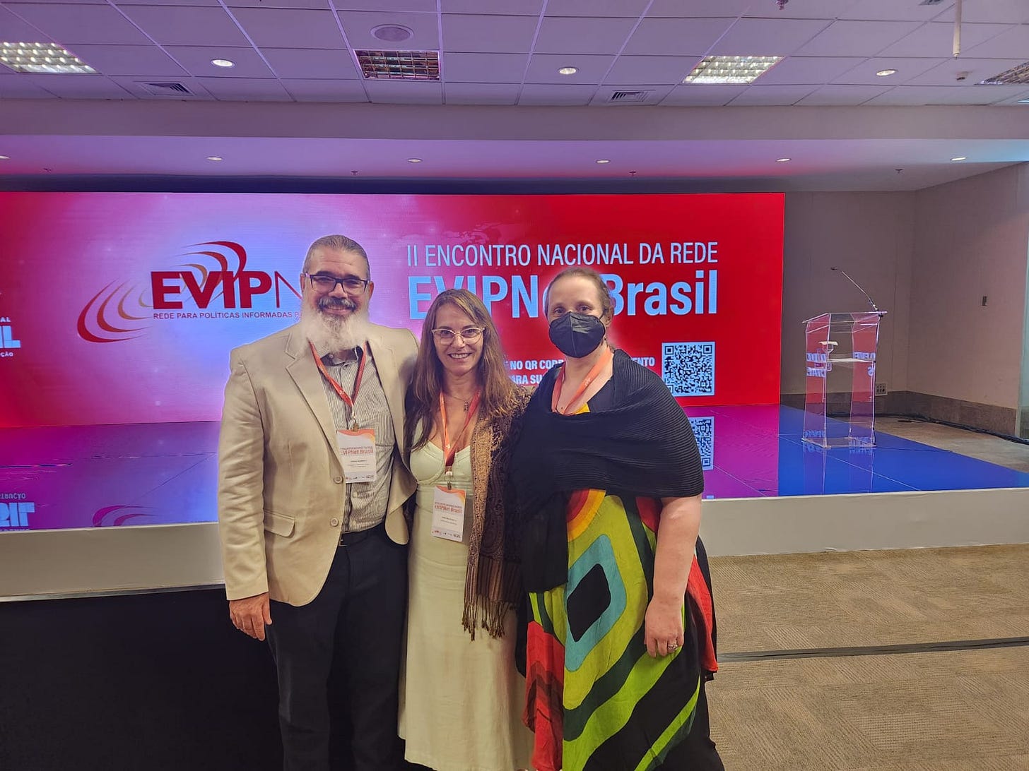 #PraTodosVerem: Da esquerda para a direita, um homem branco com cabelos grisalhos presos e barba branca. Ele usa óculos de grau, paletó bege, camisa listrada em cinza e branco, e calça social escura. Ao lado dele, Cristina Rabelais, mulher branca de cabelos castanho-claro e compridos, usa óculos de grau, um xale e vestido bege. Ambos sorriem para a foto. Em seguida, Maritsa Bortoli, mulher de cabelos loiros presos, usa máscara de proteção preta, um xale preto e um vestido estampado nas cores verde, azul, vermelho e preto. Todos usam crachás do evento. Ao fundo, há um palco e um painel nas cores vermelho e branco, com a logomarca da EVIPNet — Rede para Políticas Informadas por Evidência — e os dizeres "II Encontro Nacional da Rede EVIPNet Brasil", com um QR Code abaixo. Fim da descrição.