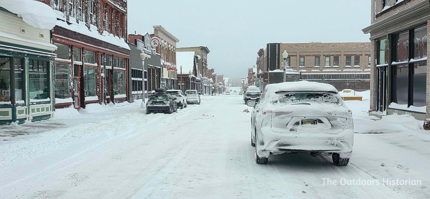 A view looking north up 5th Street in Calumet during a mid-winter snowstorm. The historic buildings are partially obscured by falling snow, and the street is nearly empty.