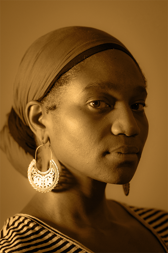 Portrait of a Black woman in warm, sepia-toned lighting with dramatic shadows. She is wearing a striped shirt, large, ornate hoop earrings, and a headscarf, and gazing directly at the camera.
