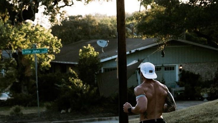 This may contain: a shirtless man running down the street in shorts and a white hat with no shirt on
