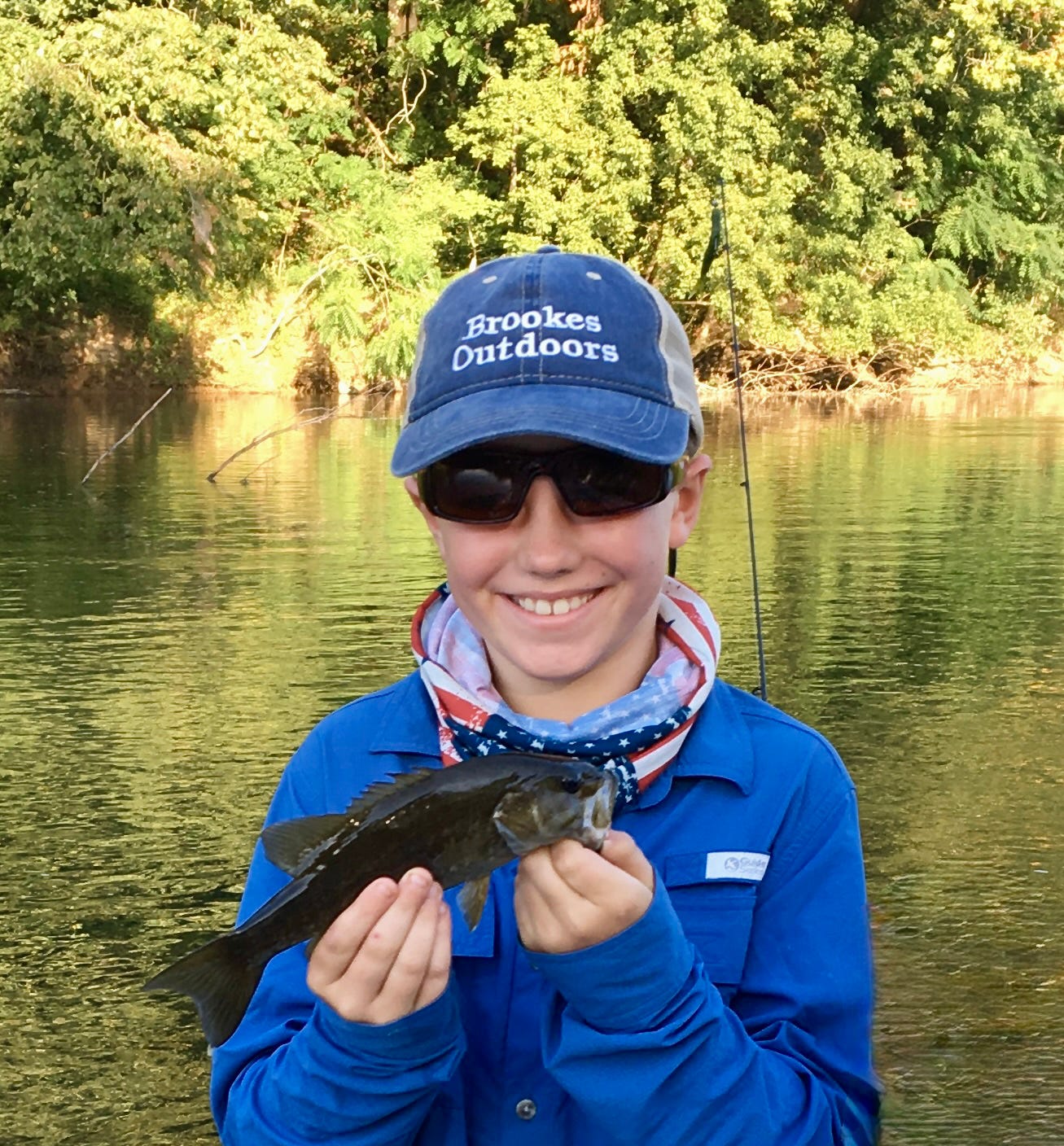 A photo of a boy holding up a smallmouth bass and smiling with a river behind him.
