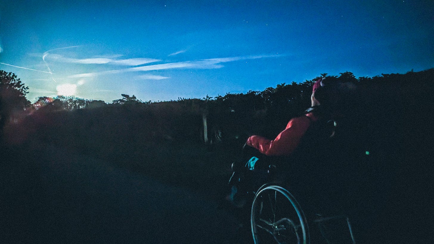 5. Landscape photograph of a night scene. The full moon is rising across the horizon. Yvie is wrapped up in thick clothing, in her wheelchair, looking out at the full moon. 5. Landscape photograph of a night scene. The full moon is rising across the horizon. Yvie is wrapped up in thick clothing, in her wheelchair, looking out at the full moon.