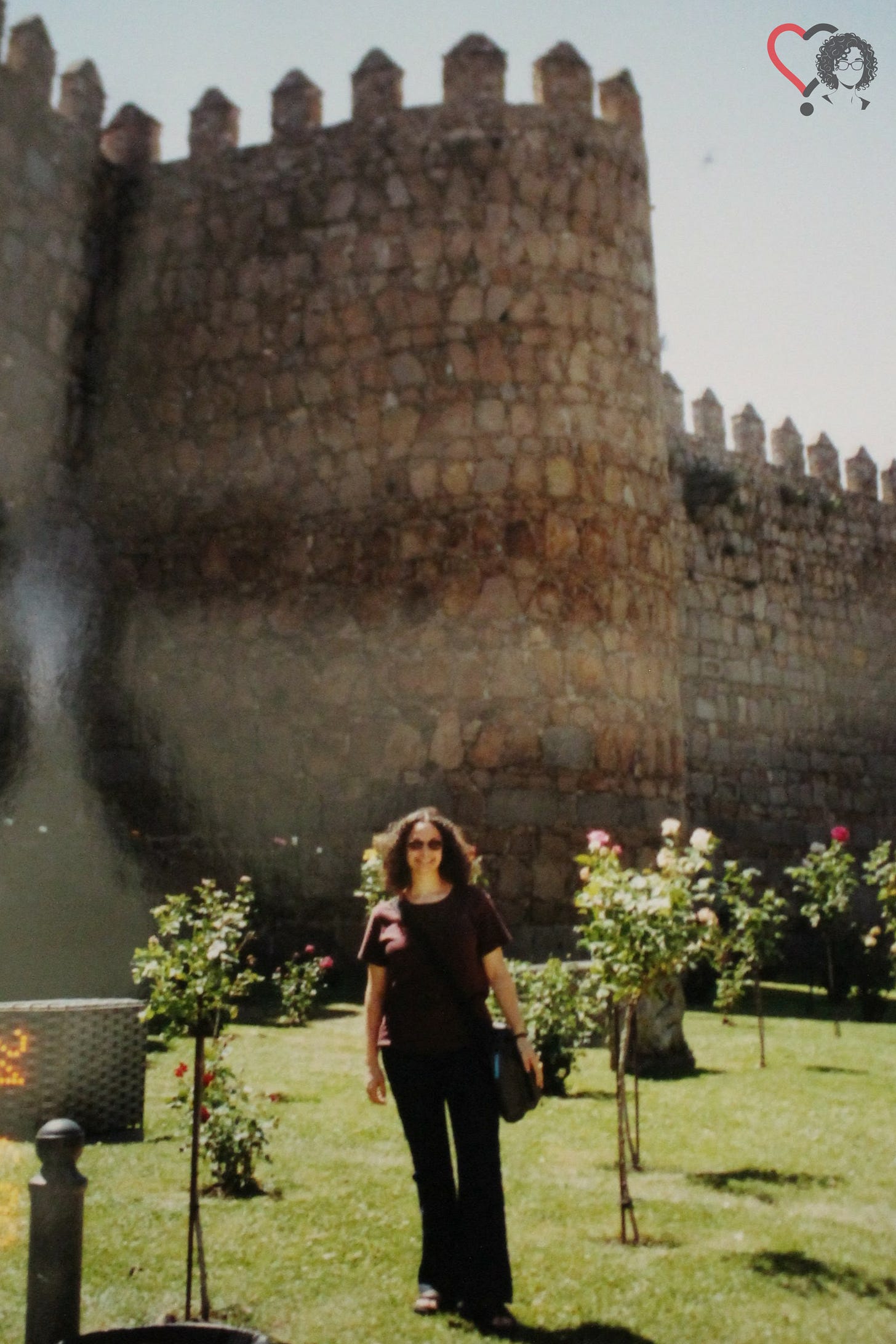 A short white woman stands in front of a Spanish Castle. She has curly, brown, shoulder-length hair and is wearing sunglasses, a burgundy t-shirt, jeans, and sandals, while carrying a black satchel on her left side. She is surrounded by small trees.