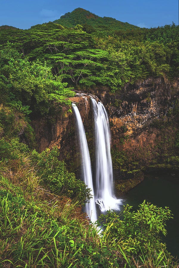 Twin Water Falls Falling off a large cliff in Hawaii and surrounded by tropical trees.