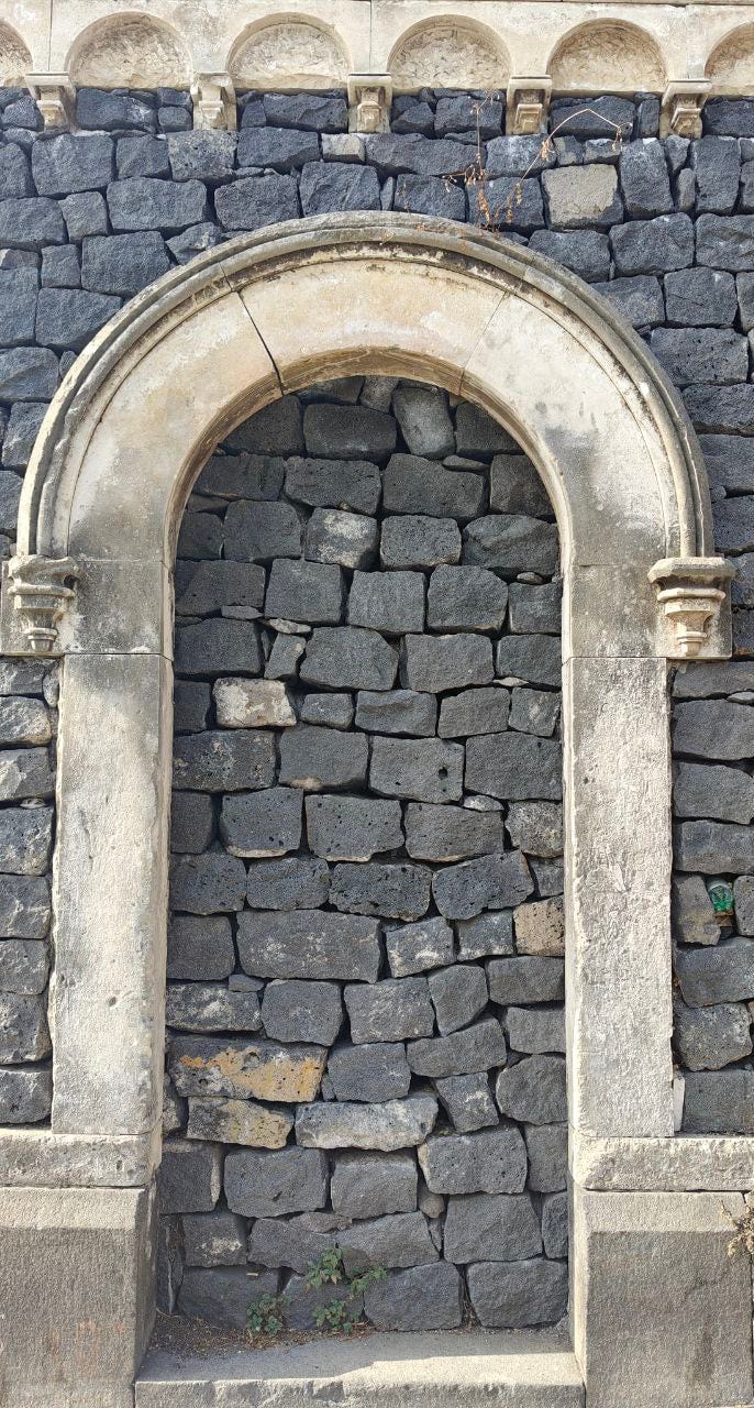Una nicchia o porta ad arco murata in una parete esterna. La parete è costruita con pietre laviche vulcaniche scure e irregolari (probabilmente basalto) che le conferiscono una texture ruvida e un colore grigio-antracite. La cornice dell'arco è realizzata in pietra chiara, lavorata e liscia, con capitelli stilizzati sui piedritti. L'interno dell'arco è tamponato con le stesse pietre scure della parete circostante. Sulla sommità del muro si intravede un elemento decorativo a cresta, anch'esso in pietra chiara. Piccole erbe infestanti spuntano alla base dell'arco e in cima al muro.