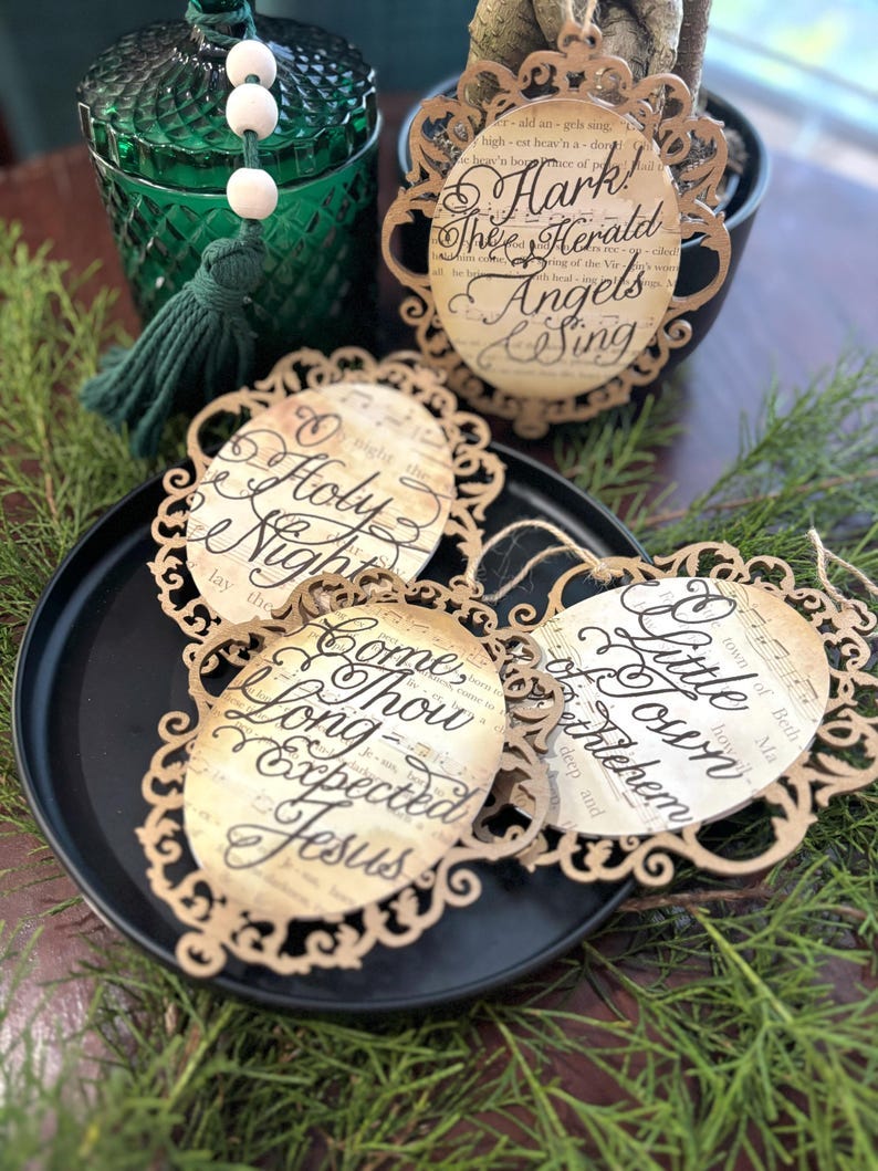 May include: Five ornate, oval-shaped Christmas ornaments. Each ornament features a different Christmas carol lyric, including 'O Holy Night,' 'Hark! The Herald Angels Sing,' 'Come Thou Long Expected Jesus,' and 'Little Town of Bethlehem.'  The ornaments are light brown with a vintage-style font and musical notes. They are arranged on a dark surface with greenery.