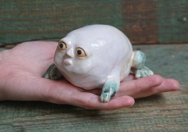 May include: A white ceramic frog figurine with green accents. The frog has a round body and large, expressive eyes. The frog is sitting on its hind legs with its front legs outstretched.