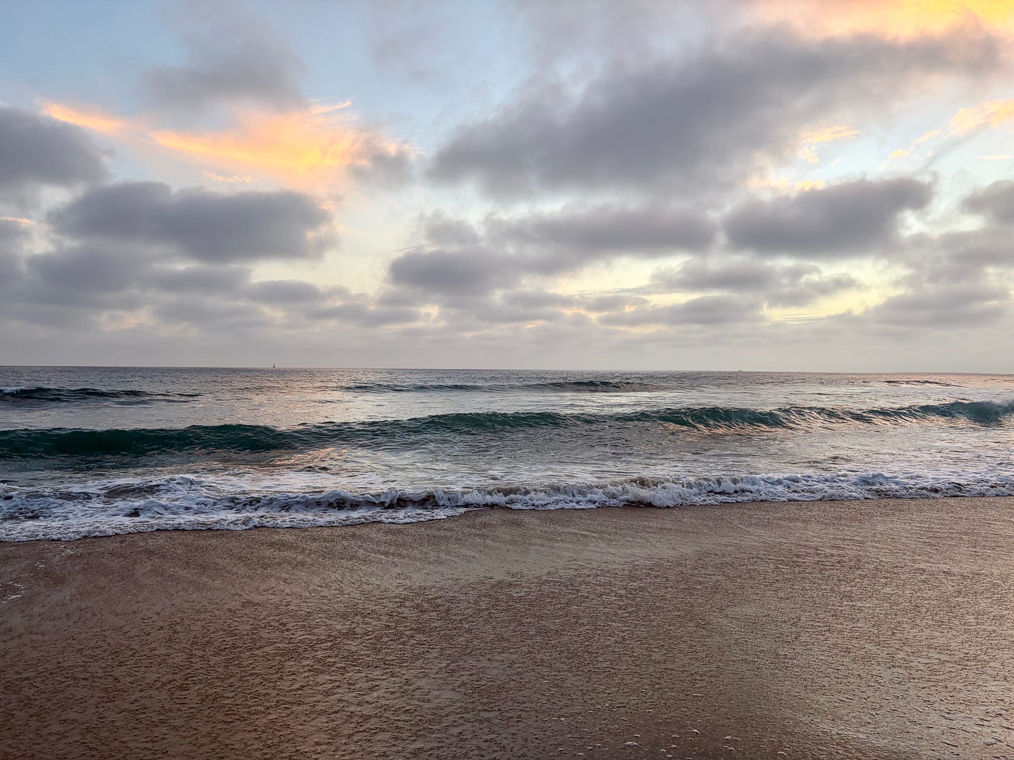 Waves rolling onto a sandy beach at dusk under a cloudy sky with soft orange light breaking through, a sailboat faint on the horizon.