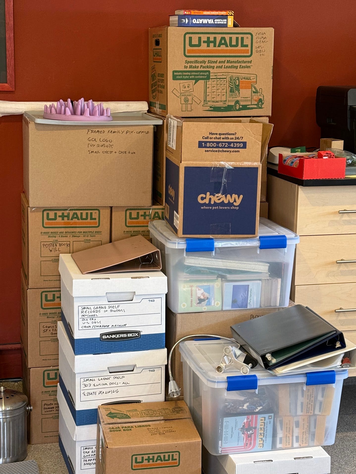 A five-high pile of Uhaul boxes and plastic bins, many labelled with a Sharpie, sitting against a red garage wall. Some contents are visible in the plastic bins, including photo albums, boxes of slides and a DVD of the documentary “Speedo.” Some binders are piled haphazardly on the boxes and bins. A cabinet of drawers is visible on the right side of the stack of boxes.
