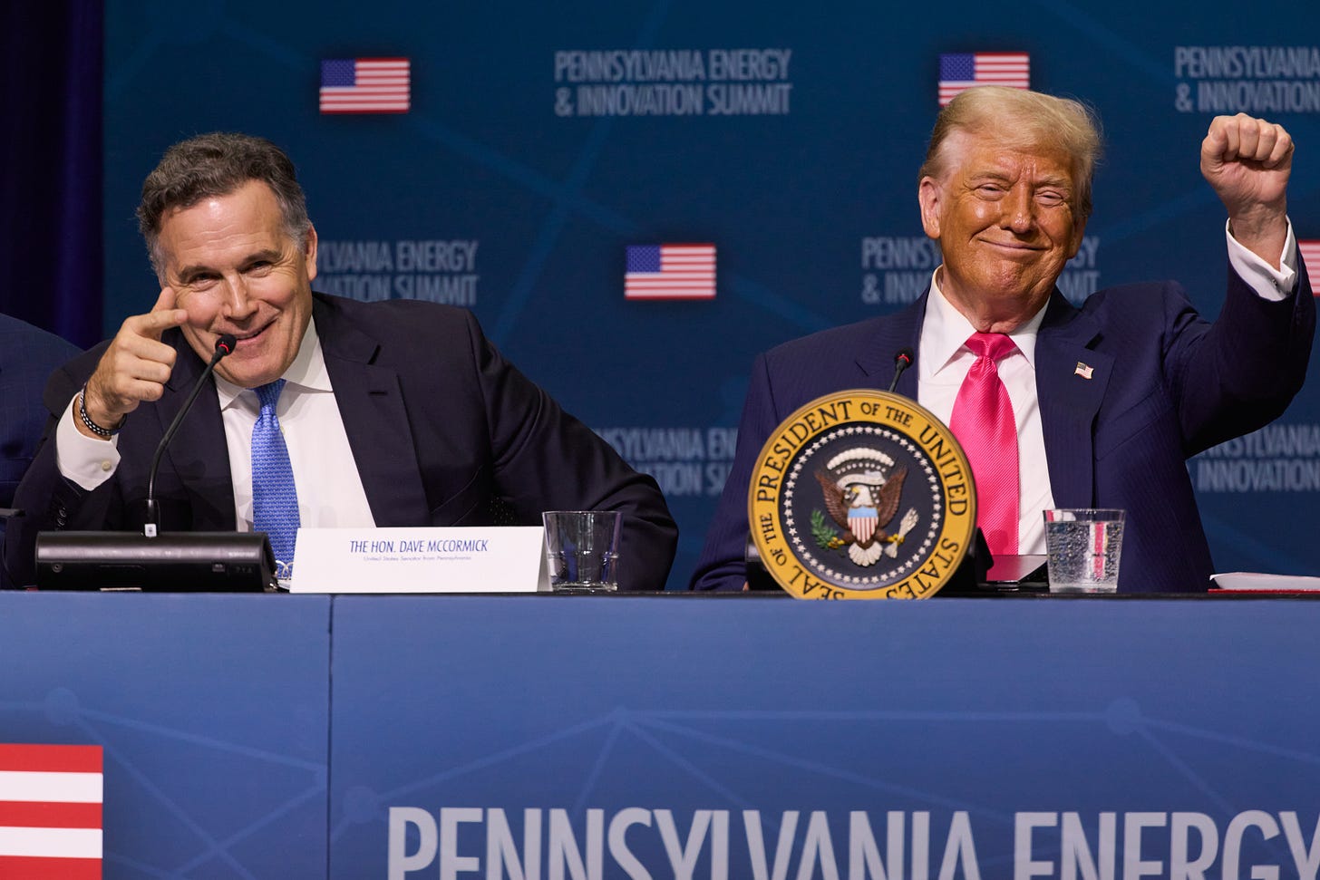 Two men in suits sit at a table. The man on the left is pointing with his index finger and the man on the right has one hand raised in a fist.