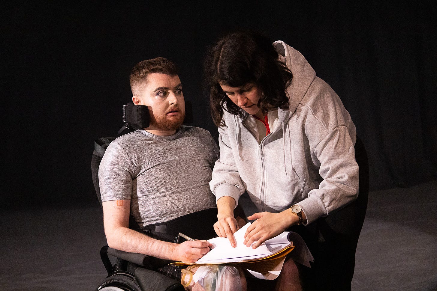 Image: Jamie Hale, a white person with dark red hair and a ginger beard sits strapped tightly into a manual wheelchair. A white woman with dark hair in a grey jumper leans over them, turning pages on a clipboard placed on Jamie’s lap, while Jamie holds a pen.
