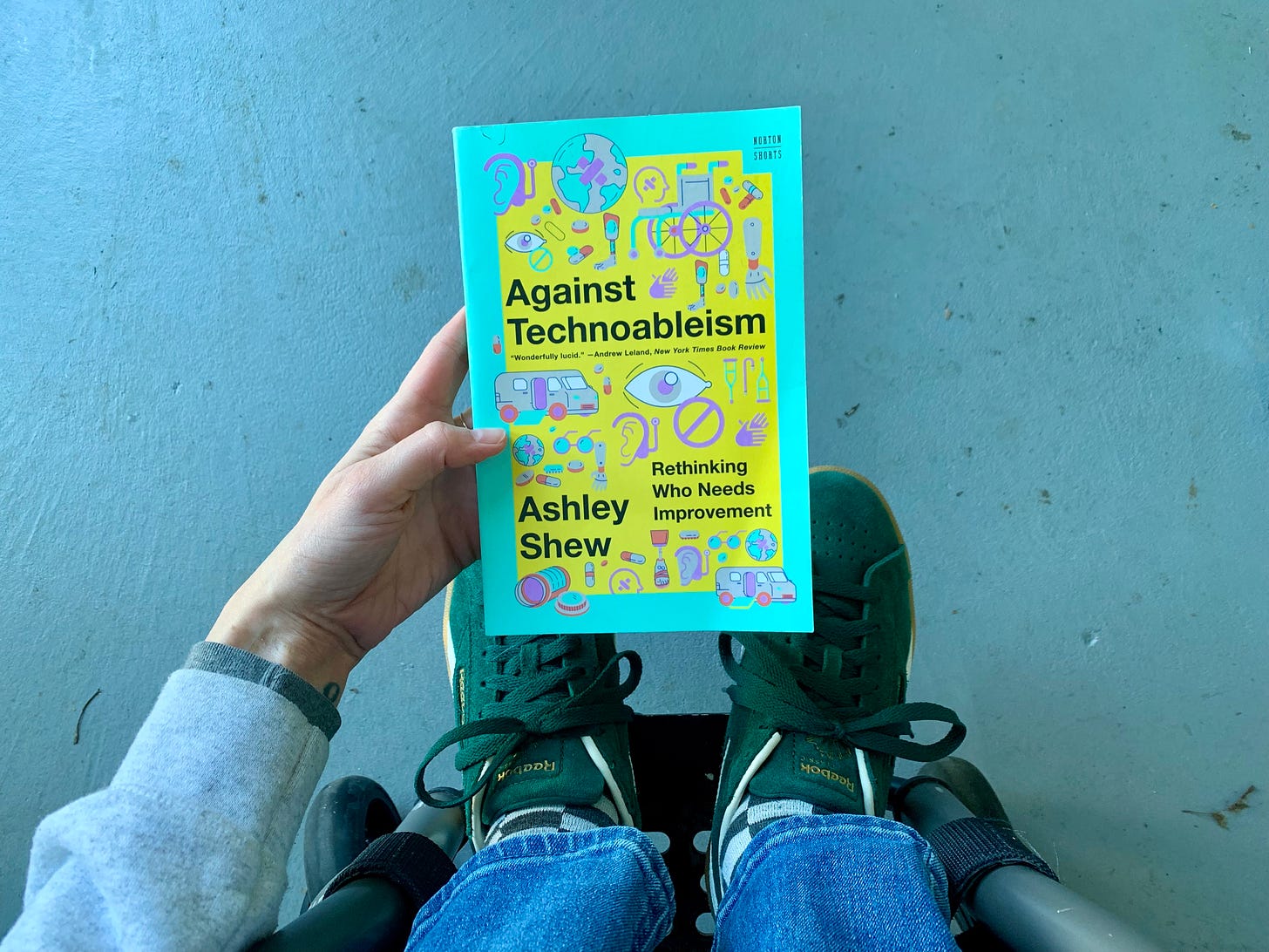 Photo taken from above, Rebekah holds a copy of Against Technoableism by Ashley Shew. The photo captures the tops of Rebekah’s shoes resting on the footplate of her wheelchair.