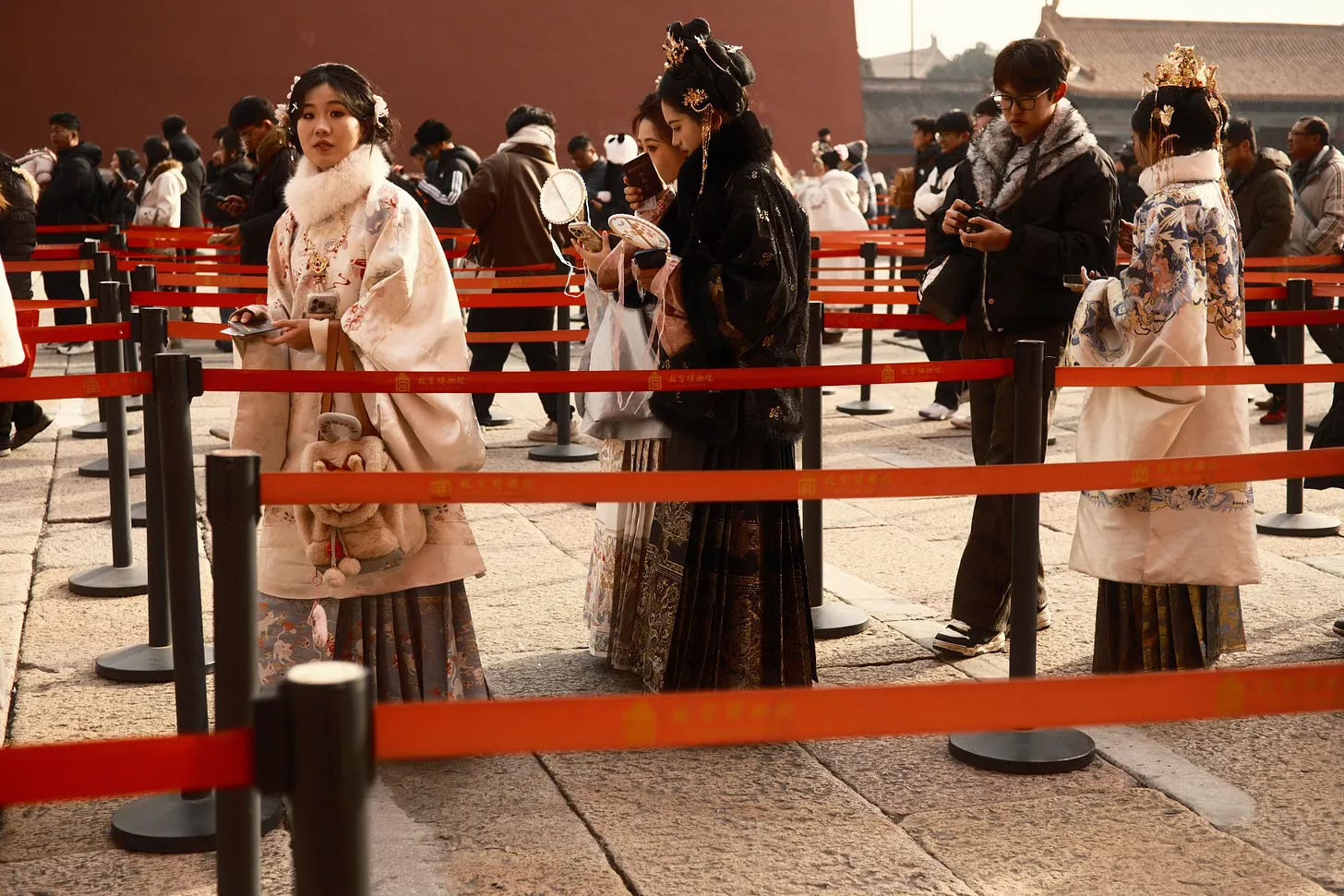 Foto van een groep bezoekers in een rij achter rode afzetlinten op een stenen plein bij de Verboden Stad in Peking. Op de voorgrond staan drie vrouwen gekleed in traditionele Chinese hanfu-gewaden: links een lichtroze ensemble met witte bontrand en goudkleurige haarversieringen, in het midden een zwart geborduurd gewaad met gouden hoofdtooi, rechts een wit-blauw gewaad met gouden kroon. Ze houden telefoons en een handspiegel vast. Op de achtergrond zijn meer bezoekers in moderne winterkleding zichtbaar. De rode afzetpaaltjes dragen Chinese tekens en een logo."