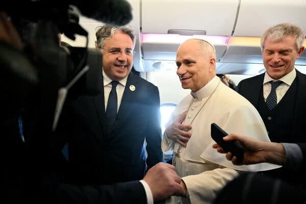 Pope Leo XIV shaking hands with someone aboard an airplane as two men in dark suits stand on either side. Pope Leo XIV shaking hands with someone aboard an airplane as two men in dark suits stand on either side.