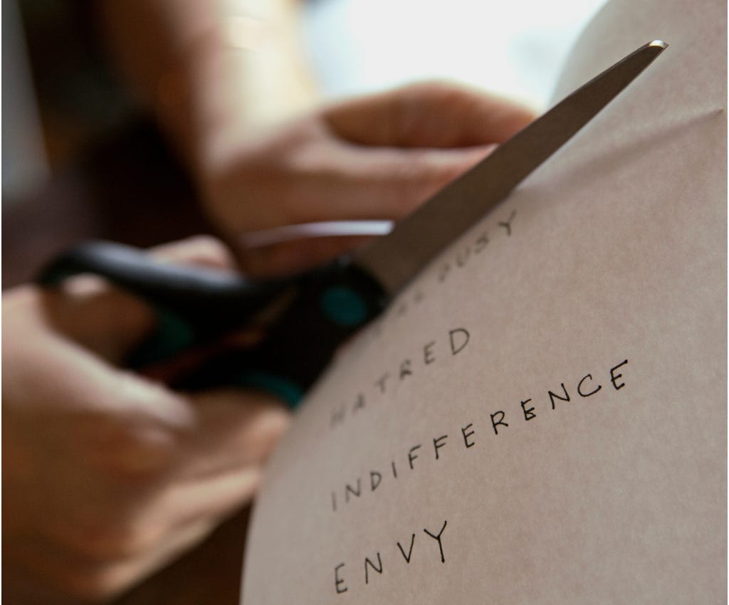 A woman’s hands are holding a sheet of white paper with a list of words on it in her right hand. The words are printed in black all-caps letters and read: too busy; hatred; indifference; envy With her left, she’s cutting these words off of the paper with stainless steel scissors with a black plastic handle. A woman’s hands are holding a sheet of white paper with a list of words on it in her right hand. The words are printed in black all-caps letters and read: too busy; hatred; indifference; envy With her left, she’s cutting these words off of the paper with stainless steel scissors with a black plastic handle.