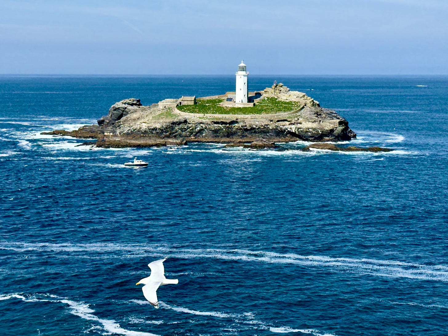 Godrevy lighthouse on its rock in St Ives Bay, a white tower above a low green plateau ringed by surf, a small boat passing and a gull crossing the foreground. Godrevy lighthouse on its rock in St Ives Bay, a white tower above a low green plateau ringed by surf, a small boat passing and a gull crossing the foreground.