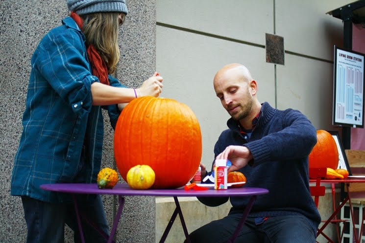 The author of the article, Max Musicant, sits at a bright purple outdoor table carving a pumpkin with a transit user at a bus station pop-up event.