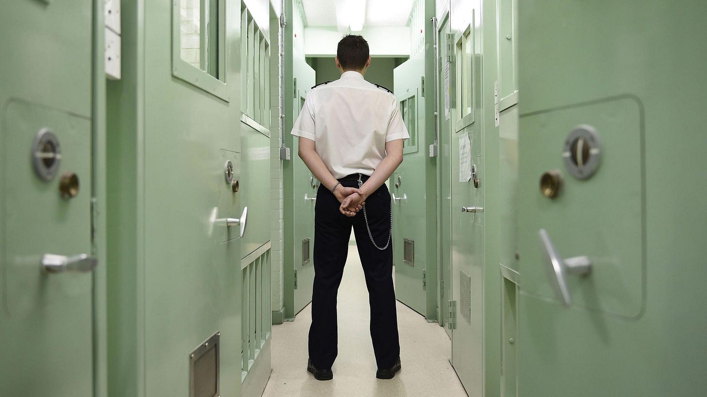 A male prison officer standing in a prison, in uniform, with his hands behind his back and his back to the camera. He's standing in a narrow corridor between two rows of cells with light green doors.